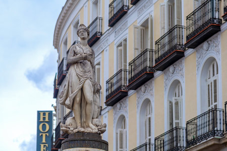 The historic statue of La Mariblanca on a tall inscribed column stands near Puerta del Sol, set against a curving, neoclassical, white-and-yellow-facade building in Madridの写真素材