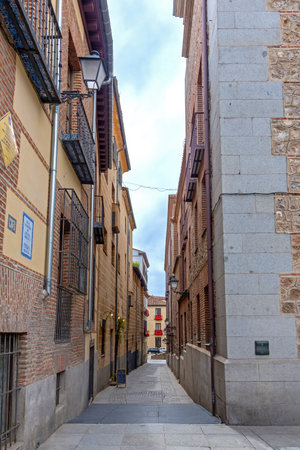 A narrow, historic alley in central Madrid, defined by towering walls of brick, rough-cut stone, and plaster buildings with wrought-iron balconies and traditional street lampsの写真素材