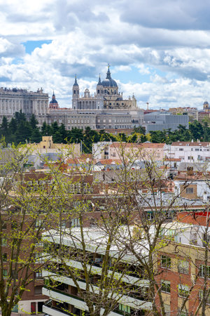 A panoramic view of the Madrid skyline shows the Royal Palace on the left and the Almudena Cathedral domes on the right, rising above lush trees and nearby residential rooftopsの写真素材