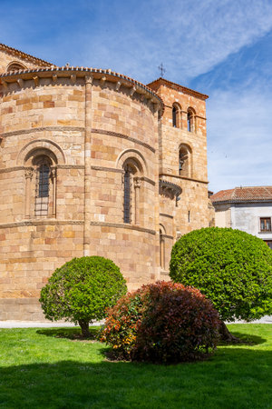 The Romanesque apse and bell tower of Ãvilaâs Church of San Pedro, built of warm golden stone, rise behind manicured green bushes and tall trees beneath a bright blue skyの写真素材