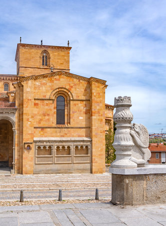 A gray stone sculpture shaped like a chess piece and lion stands before Ãvilaâs BasÃ­lica de San Vicente, a Romanesque church with a golden-brown facade and archesの写真素材