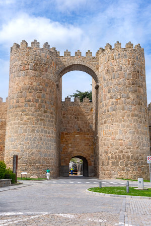The Puerta de San Vicente, a main gateway to the Walls of Ãvila, features two massive crenellated stone towers linked by an archway above a paved road and green median.の写真素材