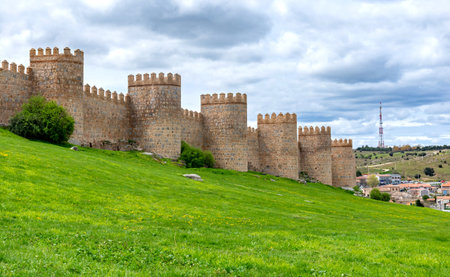 A portion of Ãvilaâs ancient stone walls, with tall rounded towers rising above a bright green embankment and set beneath a softly clouded sky that creates a calm atmosphereの写真素材
