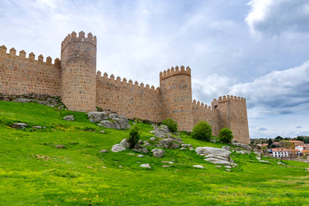 A portion of Ãvilaâs ancient stone walls, with tall rounded towers rising above a bright green embankment and set beneath a softly clouded sky that creates a calm atmosphereの写真素材