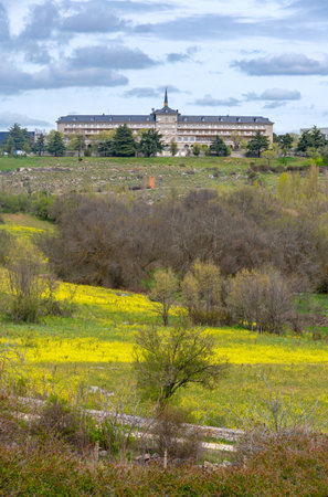 Dramatic view across a broad field of vibrant yellow wildflowers and trees toward the distant Catholic University of Ãvila, perched on a grassy hill beneath a cloudy skyのeditorial素材