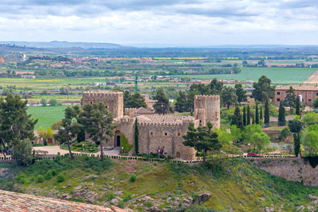 A wide view of Toledoâs Castle of San Servando, with round towers and crenellated stone walls rising among trees on a hill, overlooking the plains under a cloudy skyの写真素材