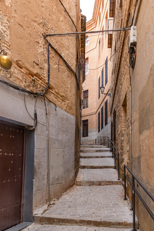 A vertical view down a narrow, winding cobblestone street in Toledo, framed by tall historic buildings with wrought-iron balconies under a cloudy skyのeditorial素材