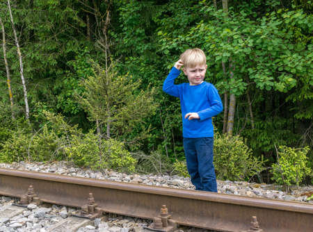 Boy caucasoid in blue clothes it stands near rails of old railway against background of forest. Emotion on his face is perplexityの写真素材