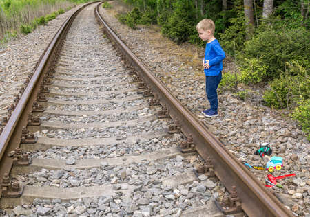 Boy caucasoid in blue clothes looks at the rails of the railway near forest. Emotion on his face is perplexityの写真素材