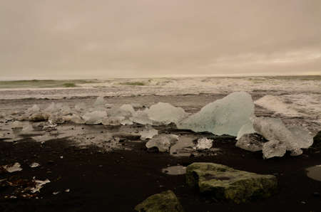A view of iceberg fragments on a black sand beach with crashing waves at the glacial lagoon in...
