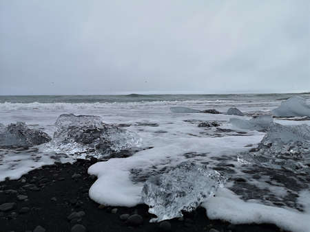 A view of iceberg fragments on a black sand beach with crashing waves at the glacial lagoon in...