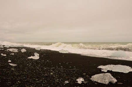 A view of iceberg fragments on a black sand beach with crashing waves at the glacial lagoon in...