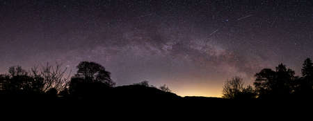 A panoramic view of the Milky Way arching over Aira Farce & the trees & mountains next to Ullswater...