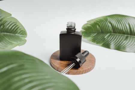 Black cosmetic serum bottle on wooden placemat. Minimalistic white background and foliageの写真素材