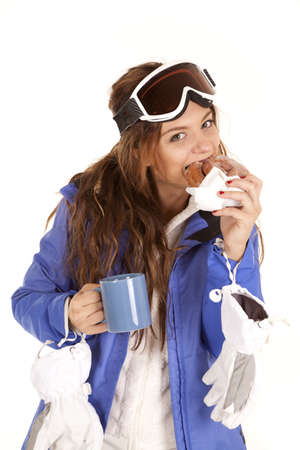 A girl in her ski out fit and goggles taking a bite of a doughnut holding her mug with a  warm drink.の写真素材