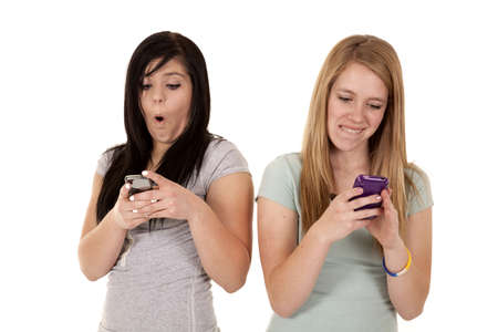two teen girls texting on their phones on has a small smile on her face the other is shocked at the text she just got.の写真素材