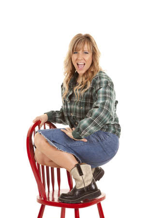 A woman in a skirt and checkered shirt, and boots on top of a red chair with a big smile on her faceの写真素材