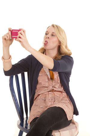 a woman sitting in her chair taking a picture of herself with her cell phone, while she sticks out her tongue.の写真素材