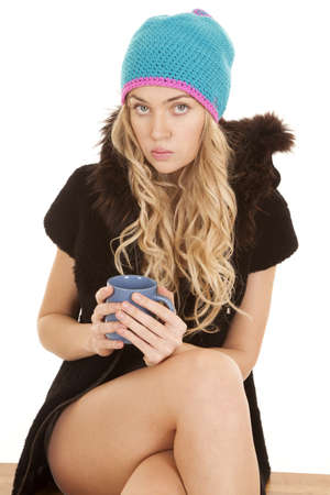 A woman with a serious expression sitting on a bench holding on to her mug in her fur jacket and warm hat.の写真素材