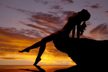 A woman is sitting in the sunset silhouetted on a rock by the water.の写真素材