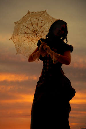 A woman in the beautiful orange sunset in her vintage dress holding on to her umbrella.の写真素材
