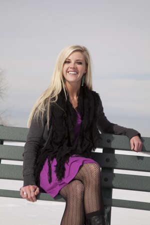A woman sitting on a green park bench in the outdoors in the snow with a smile on her face.の写真素材