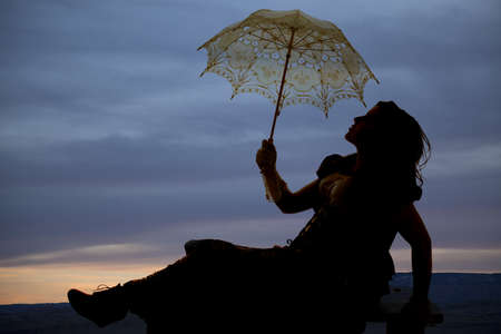 a silhouette of a woman sitting on a hill holding on to her umbrella with the beautiful sky behind her.の写真素材