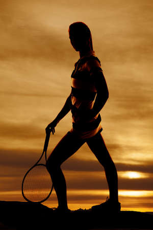 A silhouette of a woman standing on a hill holding on to her tennis racquet with a beautiful sky behind her.の写真素材