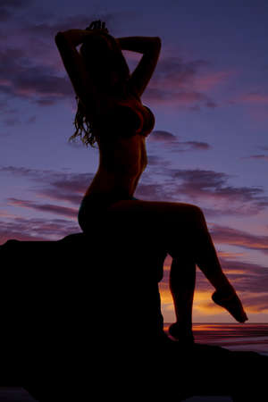 A woman in a bikini in the sunset sitting on a rock by the water.の写真素材