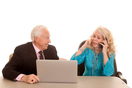a older man working on the computer while the older woman is plugging her ear so she can talk on the phone.の写真素材