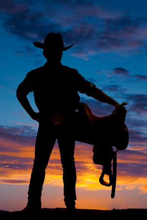 A silhouette of a man cowboy holding a saddle.の写真素材
