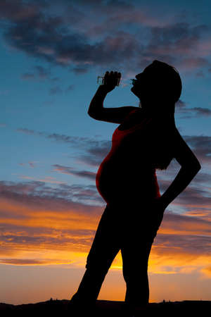 a silhouette of a pregnant woman drinking a water bottle in the outdoors.の写真素材