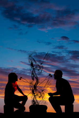 A silhouette of a couple eating and roasting marshmallows over a camp fire.の写真素材