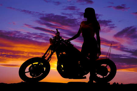 A silhouette of a woman standing next to a motorcycle looking to the side.の写真素材