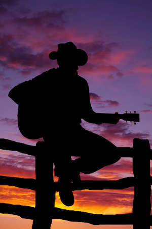 A silhouette of a cowboy sitting on a fence playing his guitar.の写真素材