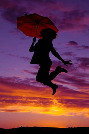 A silhouette of a woman jumping up in her rain boots, and holding an umbrella.の写真素材