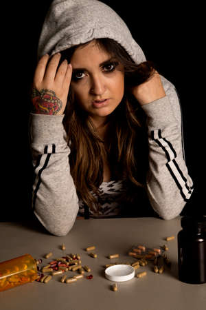 A woman with her jacket pulled over her head, looking at the pills on the table, with a troubled expression on her face.の写真素材