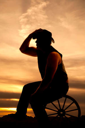 A silhouette of a cowboy sitting on a wagon wheel with his hand on his hat.の写真素材