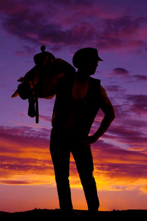 a silhouette of a cowboy with a saddle on his shoulder looking to the side.の写真素材