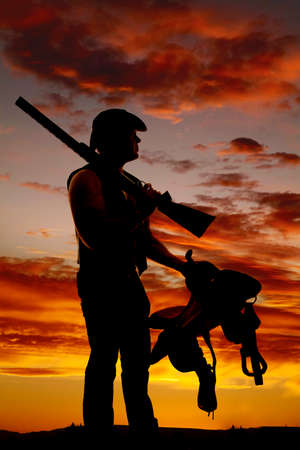 A silhouette of a cowboy holding on to his saddle with a rifle on his shoulder.の写真素材