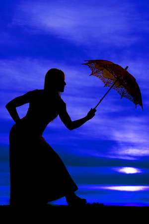 A silhouette of a woman in her dress, holding on to her umbrella, leaning over.の写真素材