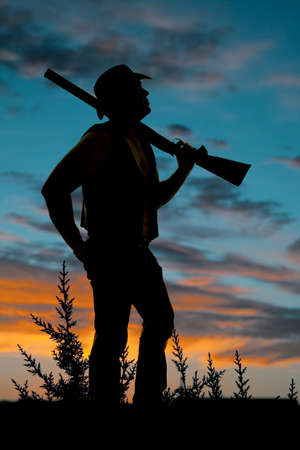 a silhouette of a cowboy standing in the weeds with a shotgun on his shoulder.の写真素材