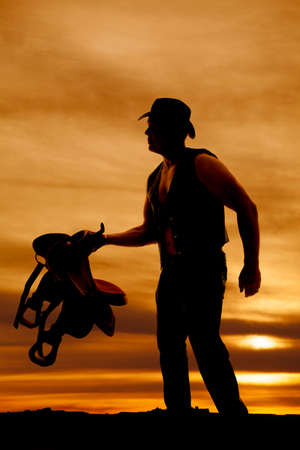 a silhouette of a cowboy holding out a saddle in the sunsetの写真素材