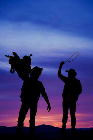 a silhouette of two cowboys, one with a saddle and the other with a rope swinging above his head.の写真素材