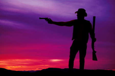 A silhouette of a cowboy pointing his pistol and holding on to his shotgun.の写真素材