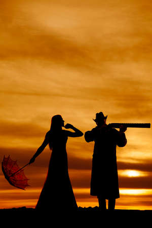 a silhouette of a woman with her umbrella looking to her cowboy who is pointing his rifle.の写真素材