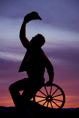 A silhouette of a cowboy sitting on a wooden wagon wheel with his hat up off of his head.の写真素材