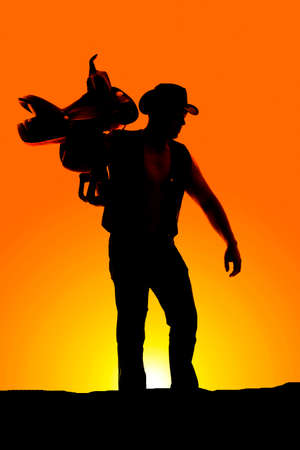 A silhouette of a cowboy with a saddle on his shoulder looking down.の写真素材