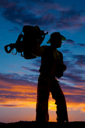a silhouette of a cowboy in the outdoors holding up his saddle, looking to the side.の写真素材