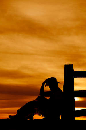 a silhouette of a cowboy sitting by a fence post touching his hat.の写真素材